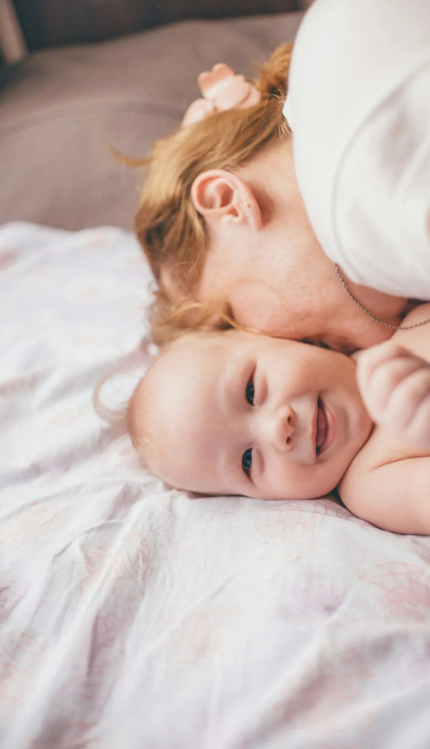 A mother lies on a bed with her cheek against her baby's head, both awake.