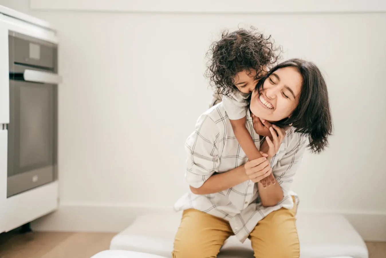 A mother sits laughing while her young child hugs her from behind, in a bright kitchen.